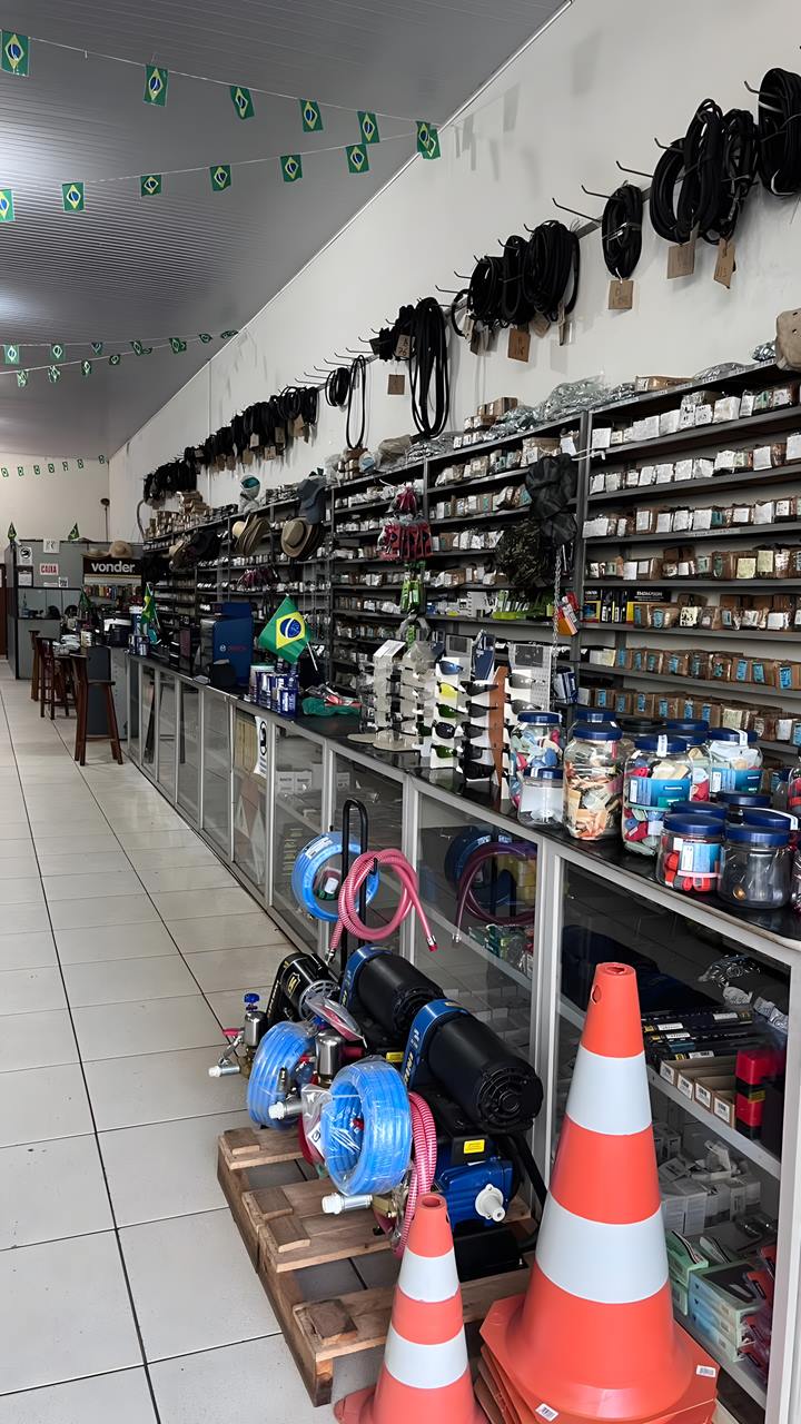 Balcão da loja com correias penduradas na parede, prateleiras cheias de caixinhas de parafuso, óculos de proteção, máquinas e cones